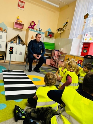 Policjant opowiadający o zasadach bezpieczeństwa ruchu drogowego. Wokoło dzieci siedzące na podłodze.
